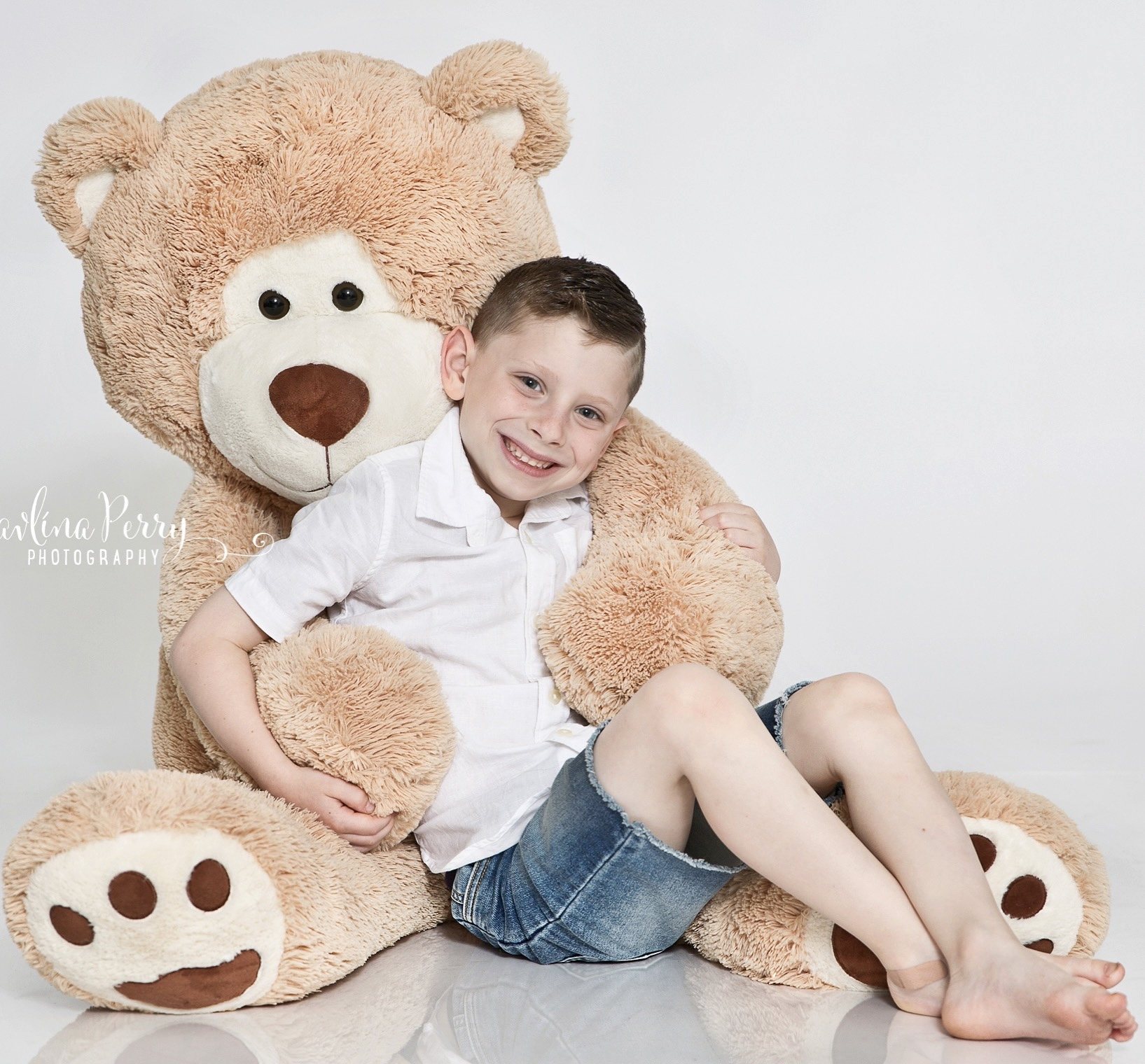 Smiling boy leaning against a large teddy bear in a bright studio