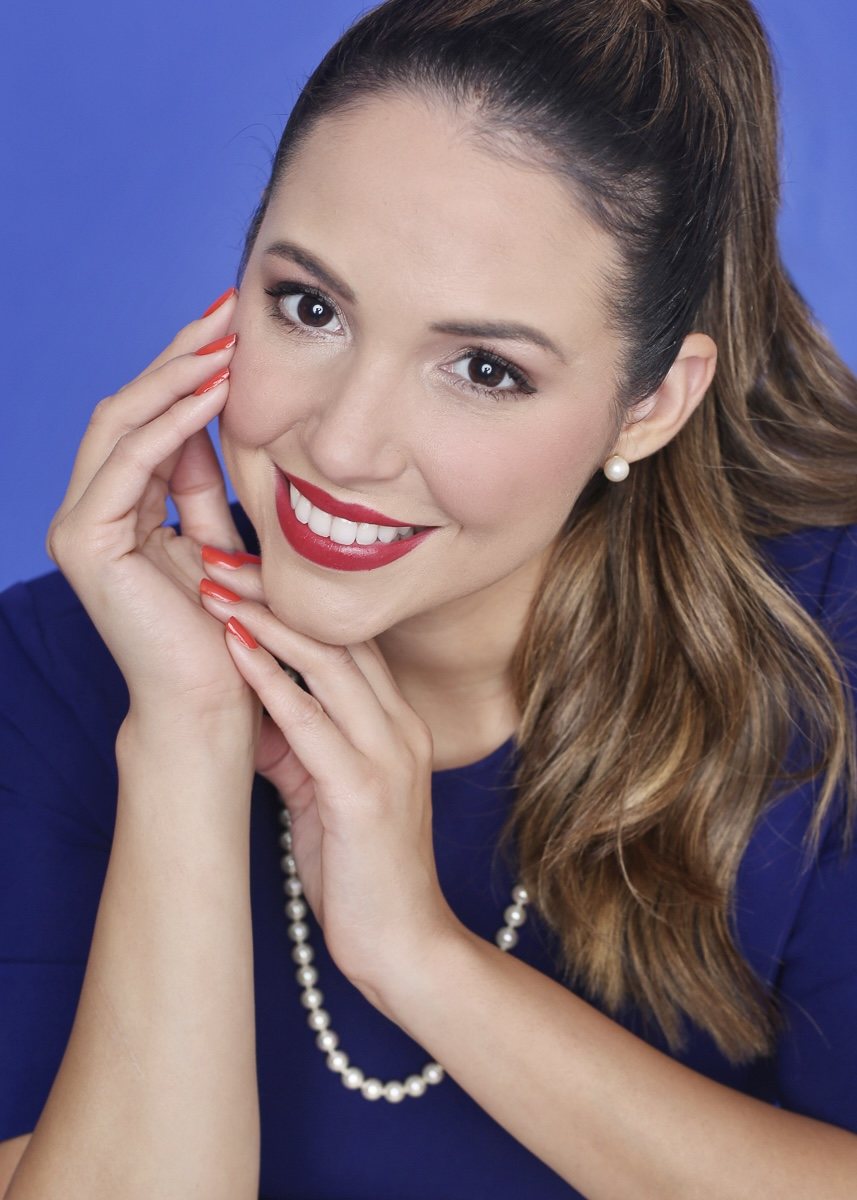 Smiling woman in blue with pearl jewelry and her hands beside her face against a blue studio background