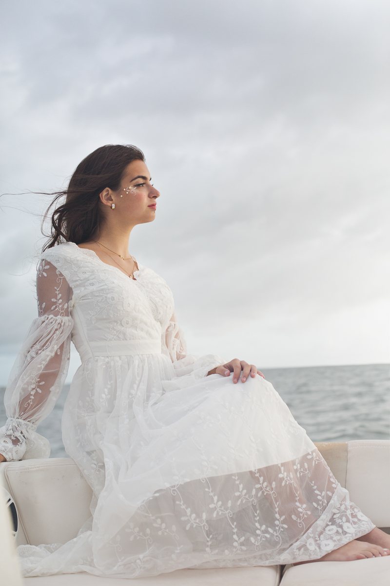 Full-length profile portrait in a flowing white lace dress, seated beside the sea with wind in her hair
