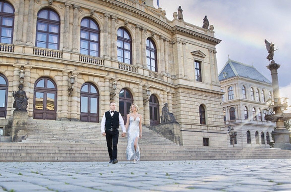 Couple holding hands in formal attire in front of grand historic architecture.