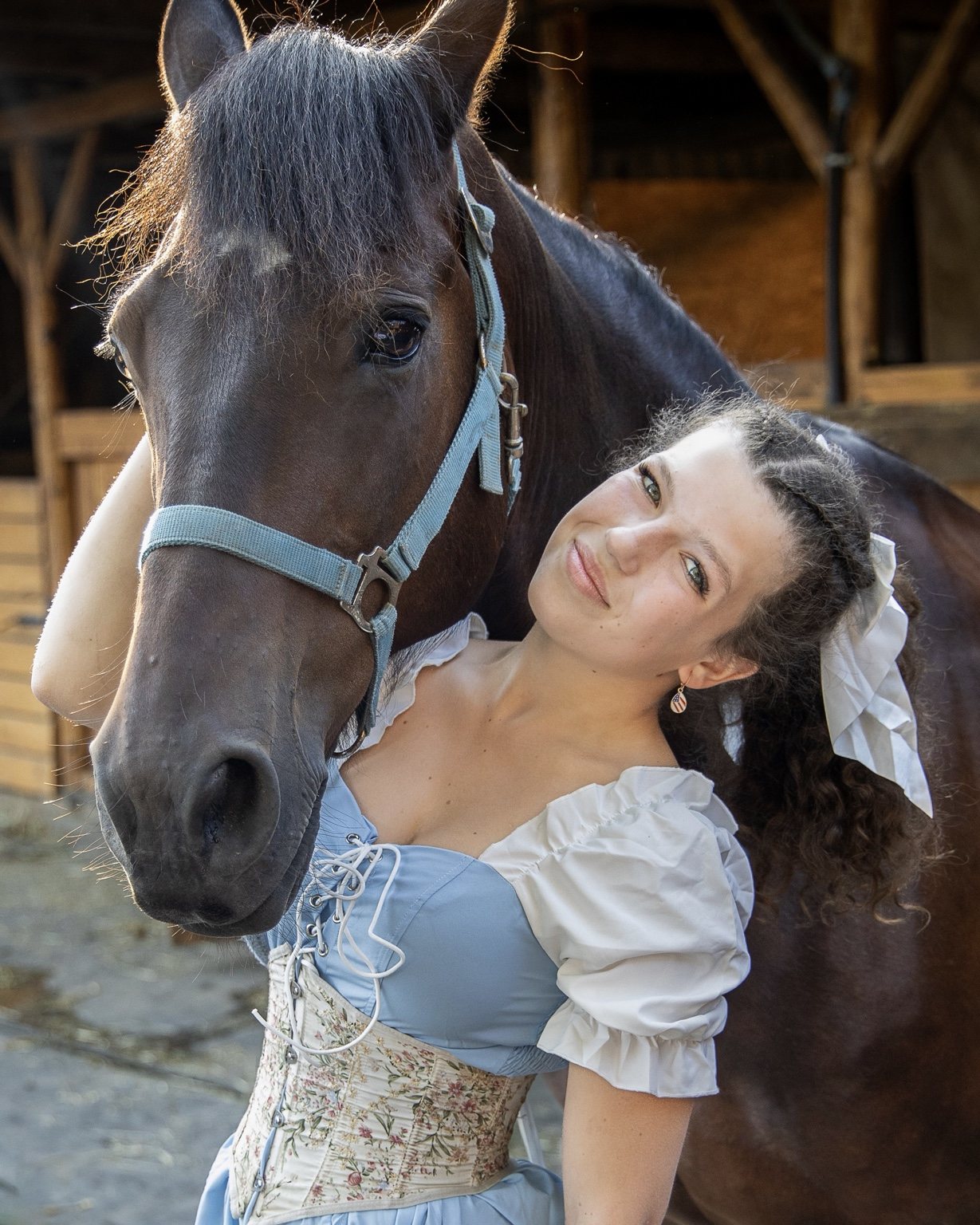 Woman in a blue dress leaning against a brown horse in a blue halter, with warm light around its mane
