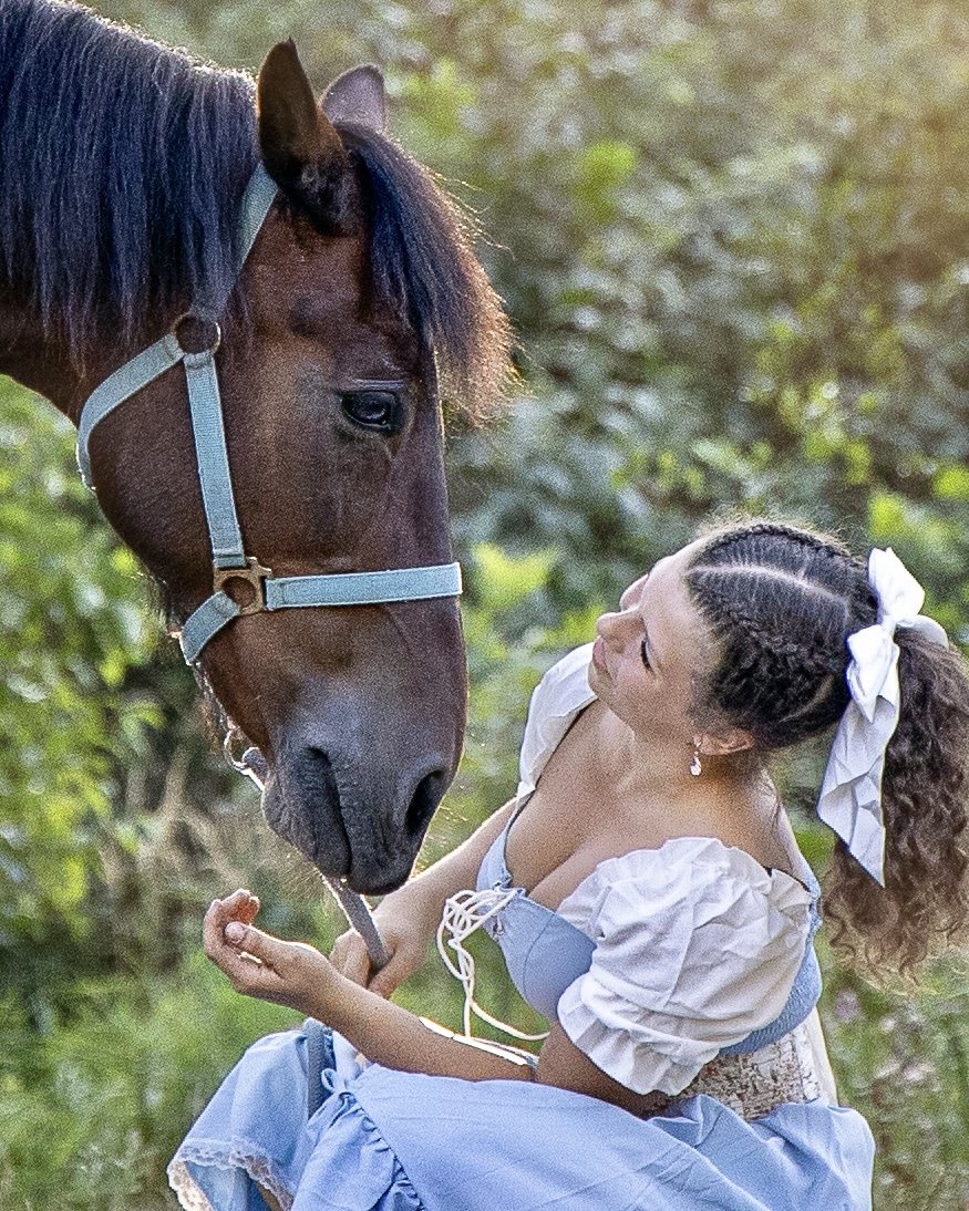 Young woman in a soft blue dress looking toward a brown horse in a blue halter, surrounded by greenery