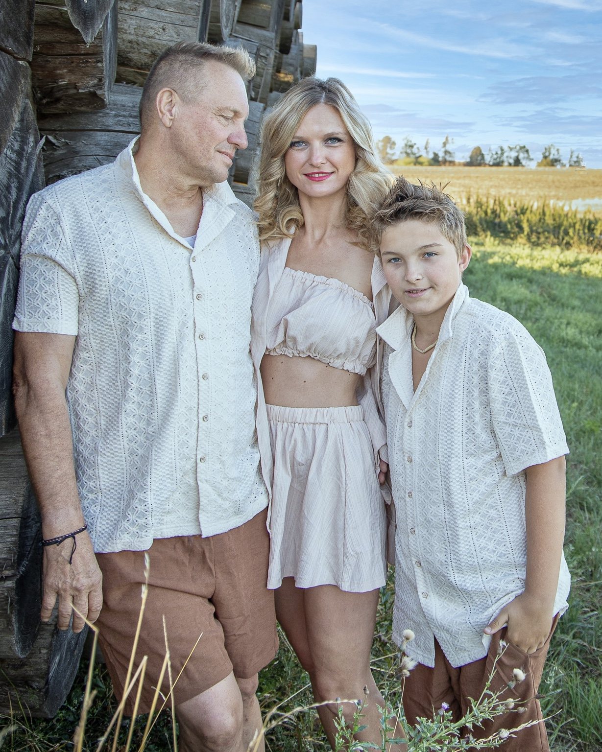 Family of three standing close beside a rustic wooden wall, with fields and morning sky behind them