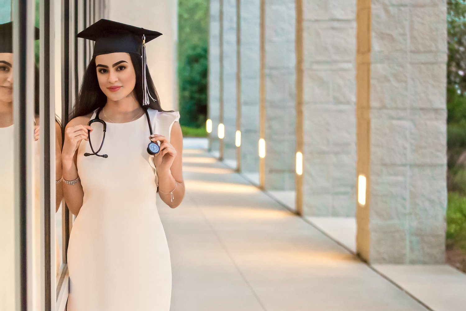 Graduate in a cap and white dress holding a stethoscope beside reflective windows