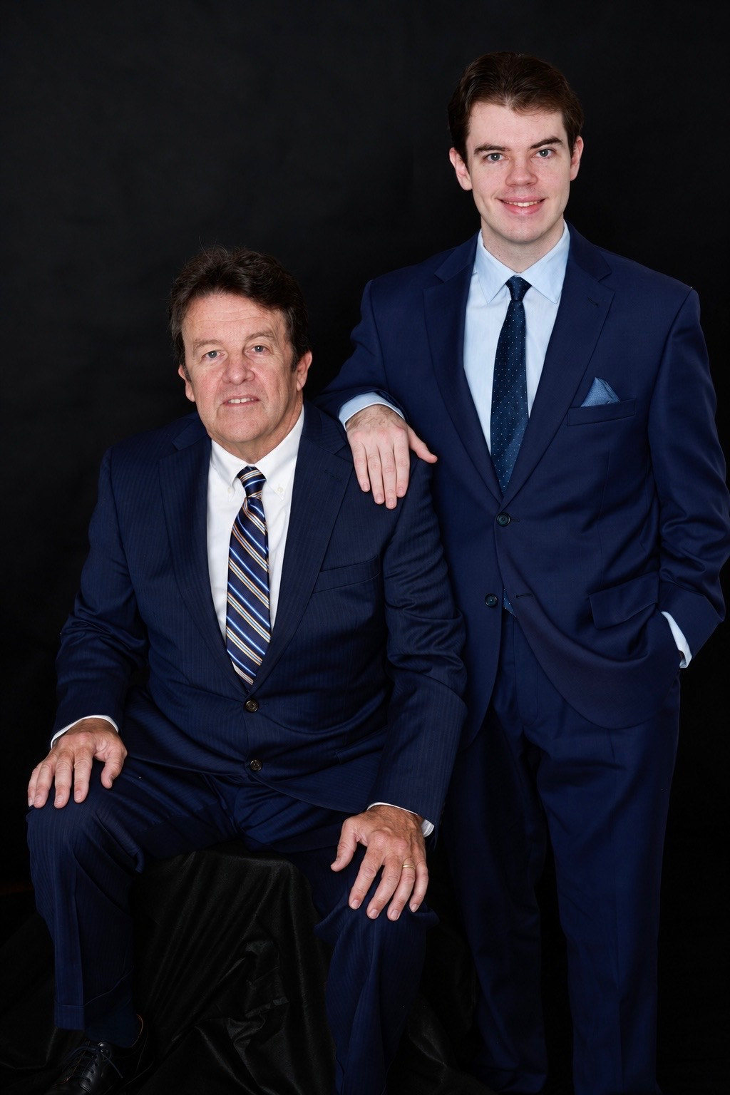 Father seated and his grown son standing beside him, both in navy suits against a black background