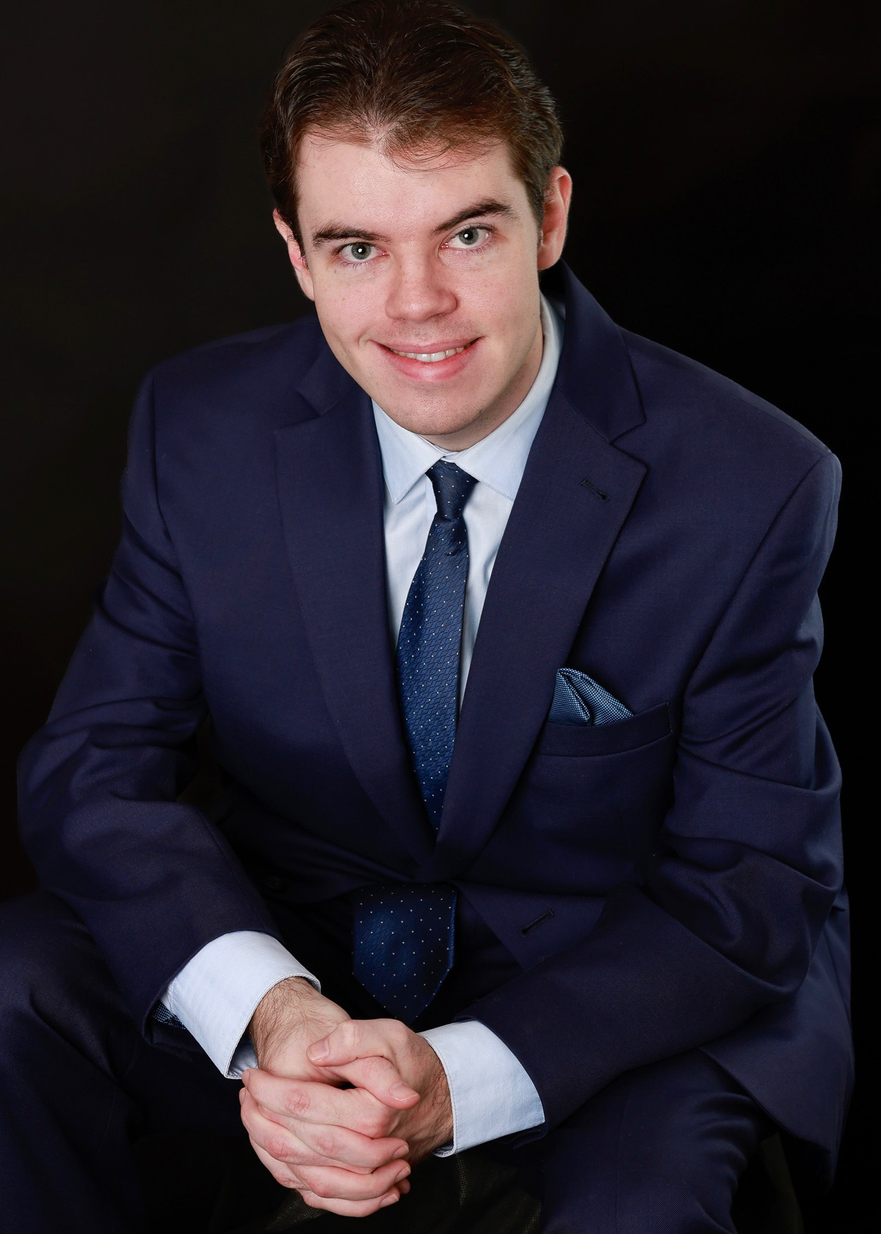 Young man in a navy suit and tie seated with hands clasped against a black background