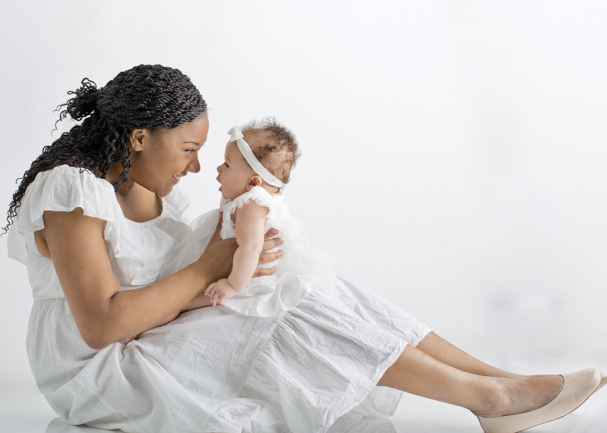 Mother in a white dress smiling at her baby, who wears a white dress and bow headband, against a light studio backdrop