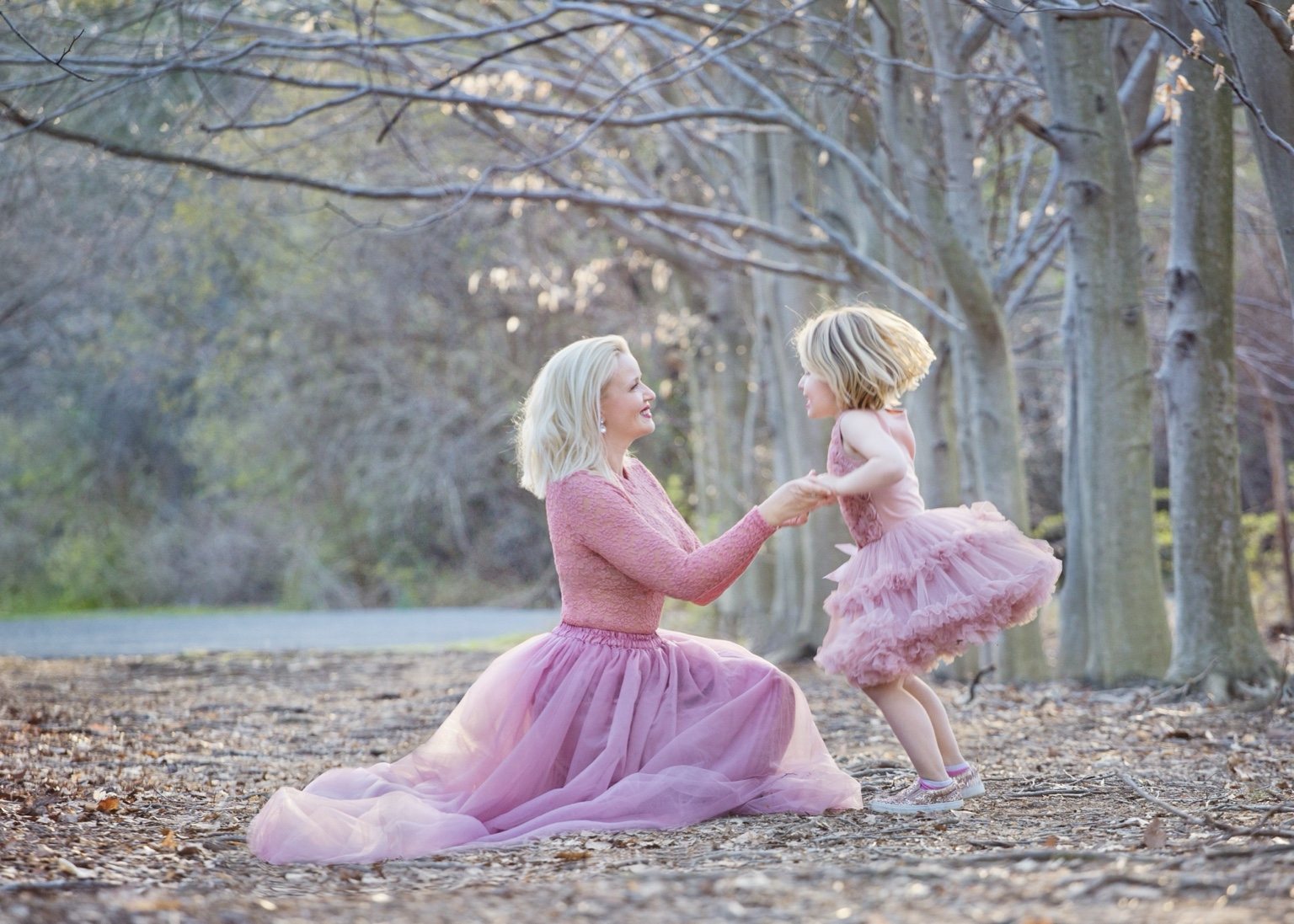 Mother and young daughter in flowing pink dresses holding hands in sunlit woodland