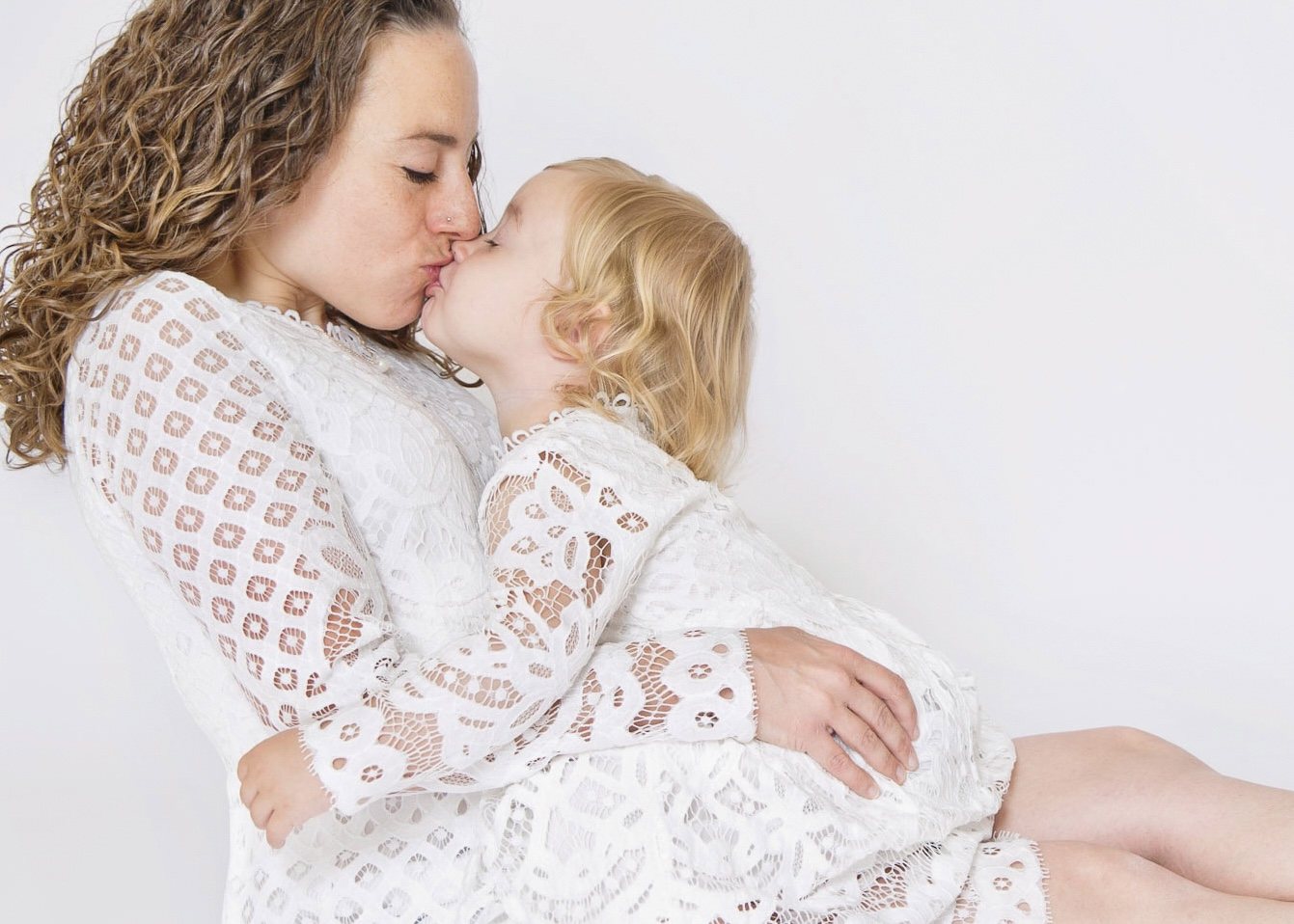 Mother and young daughter in matching white lace sharing a kiss in the studio