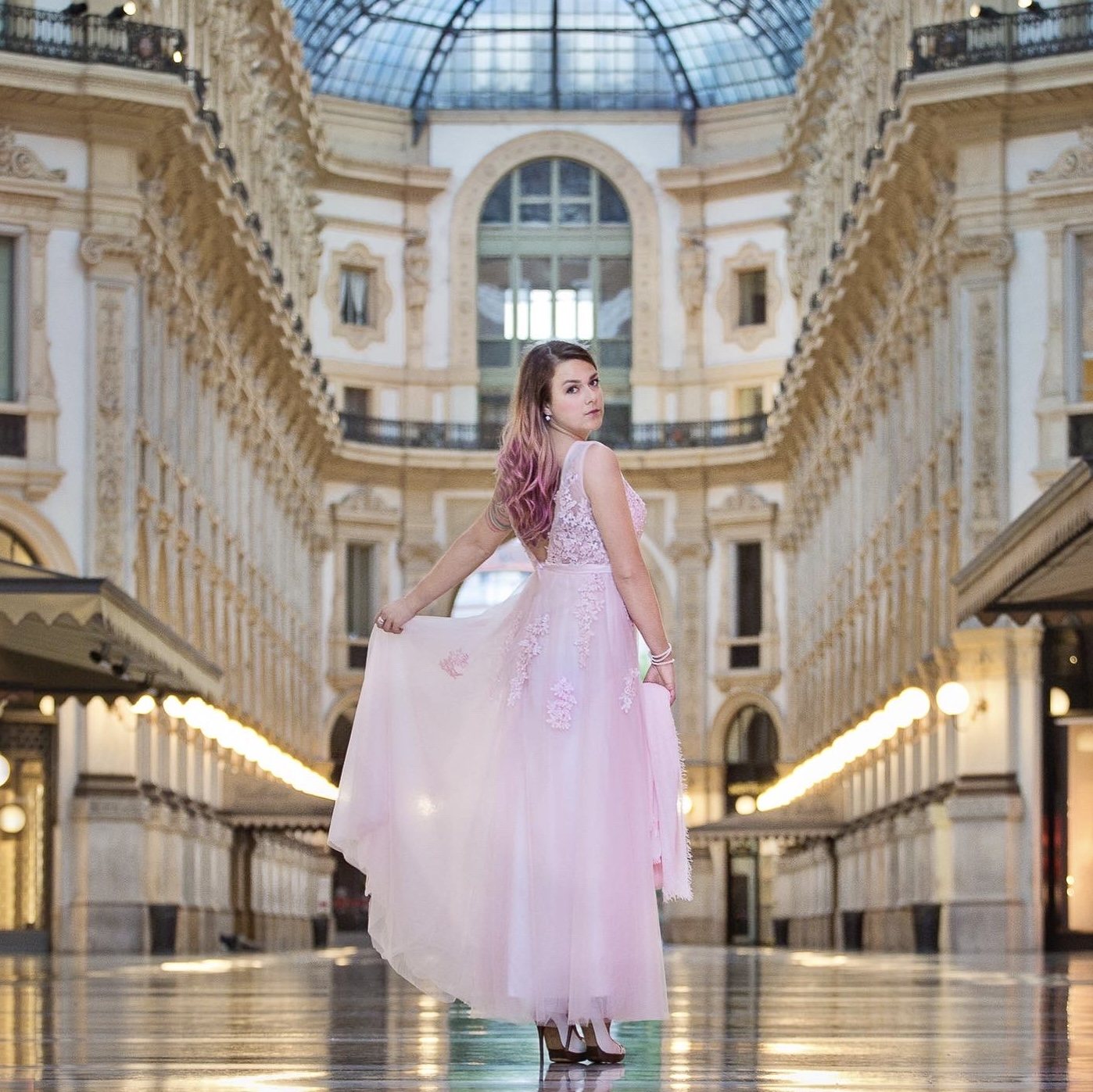 Woman in a pale pink gown looking over her shoulder beneath a glass-roofed arcade