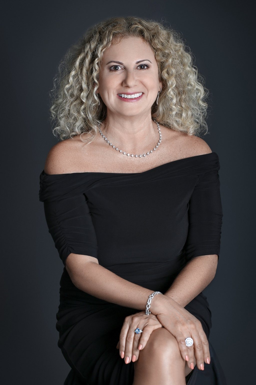 Smiling woman with curly blonde hair in a black dress against a charcoal studio background