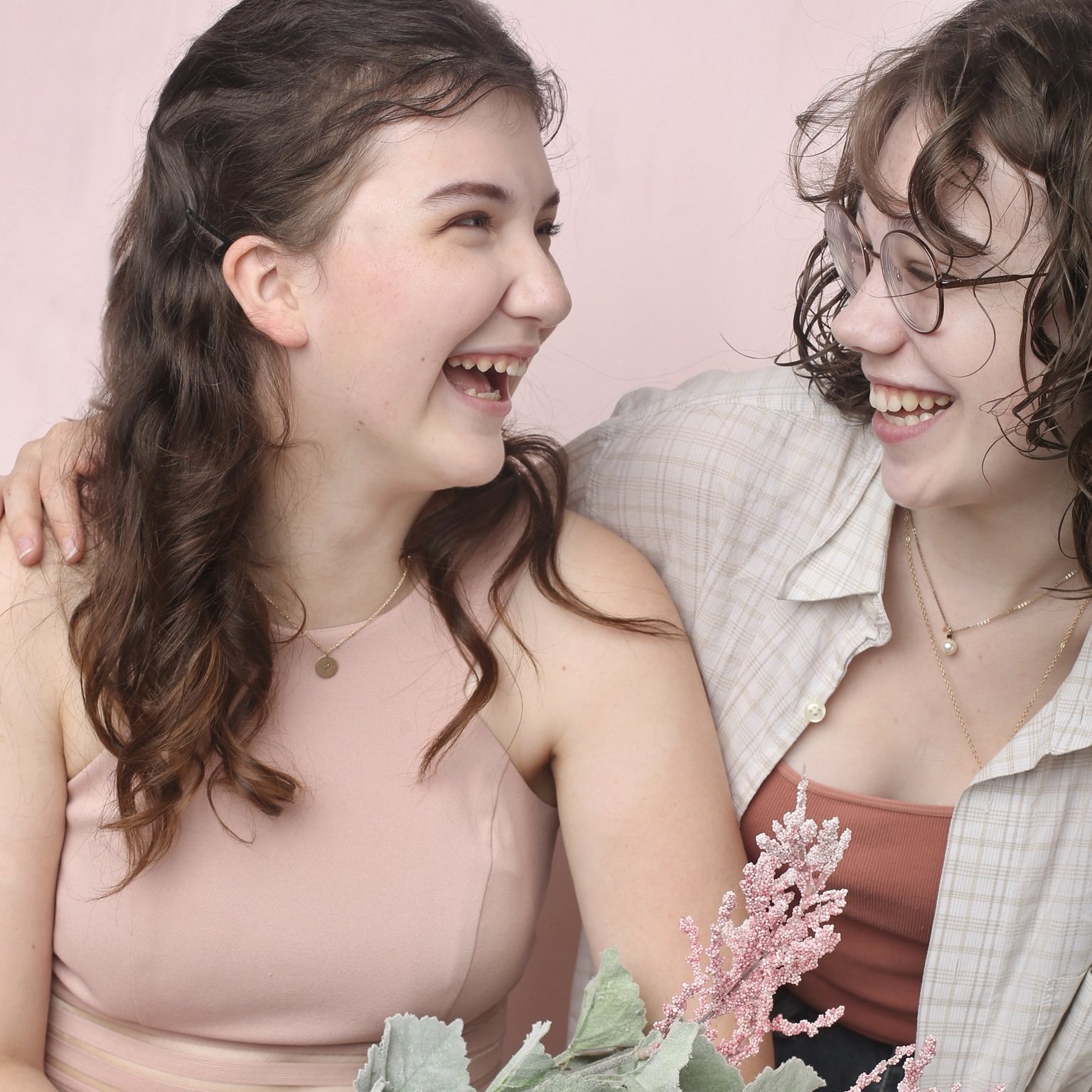 Two young women laughing together against a pale pink studio background