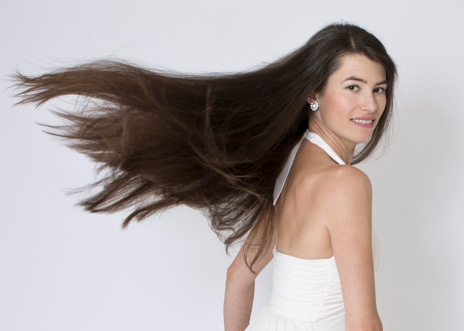 Woman in a white dress looking over her shoulder with her long brown hair flowing across a light studio background